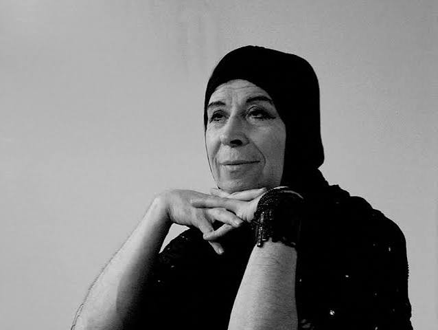 Retrato en blanco y negro de una persona con pañuelo oscuro en la cabeza y las manos apoyadas bajo el mentón.