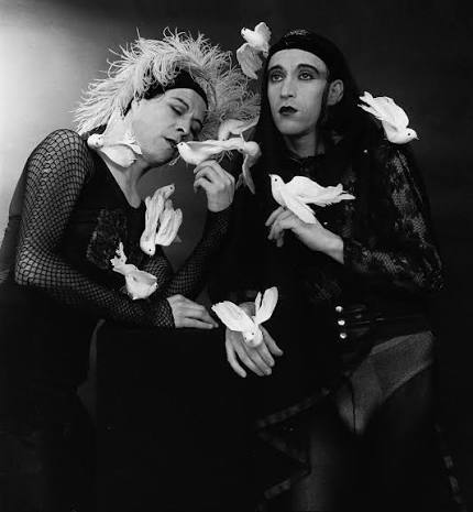 Dos artistas vestidos de negro, con maquillaje y figuras de palomas blancas, en una fotografía en blanco y negro.