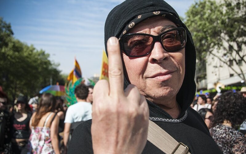 Pedro Lemebel con gorro negro y gafas de sol, levantando el dedo medio ante la cámara.