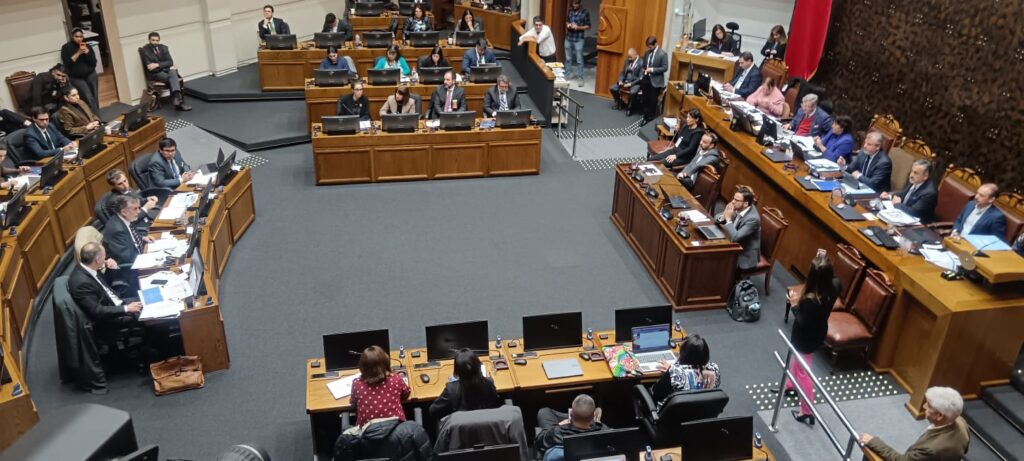 Vista general de una sesión parlamentaria, con participantes sentados ante mesas y pantallas.